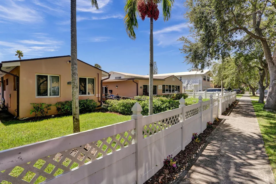Building exterior with palms, a white fence and a shaded sidewalk
